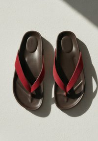 Paire de sandales marron avec de larges brides en tissu rouge projetant des ombres sur une surface claire et texturée.