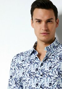 Blaues Blumen-Hemd mit Knöpfen, das ein lebhaftes Blatt- und Vogelmuster zeigt, mit einem Kragen und unterschiedlichen Blautönen auf einem weißen Hintergrund.