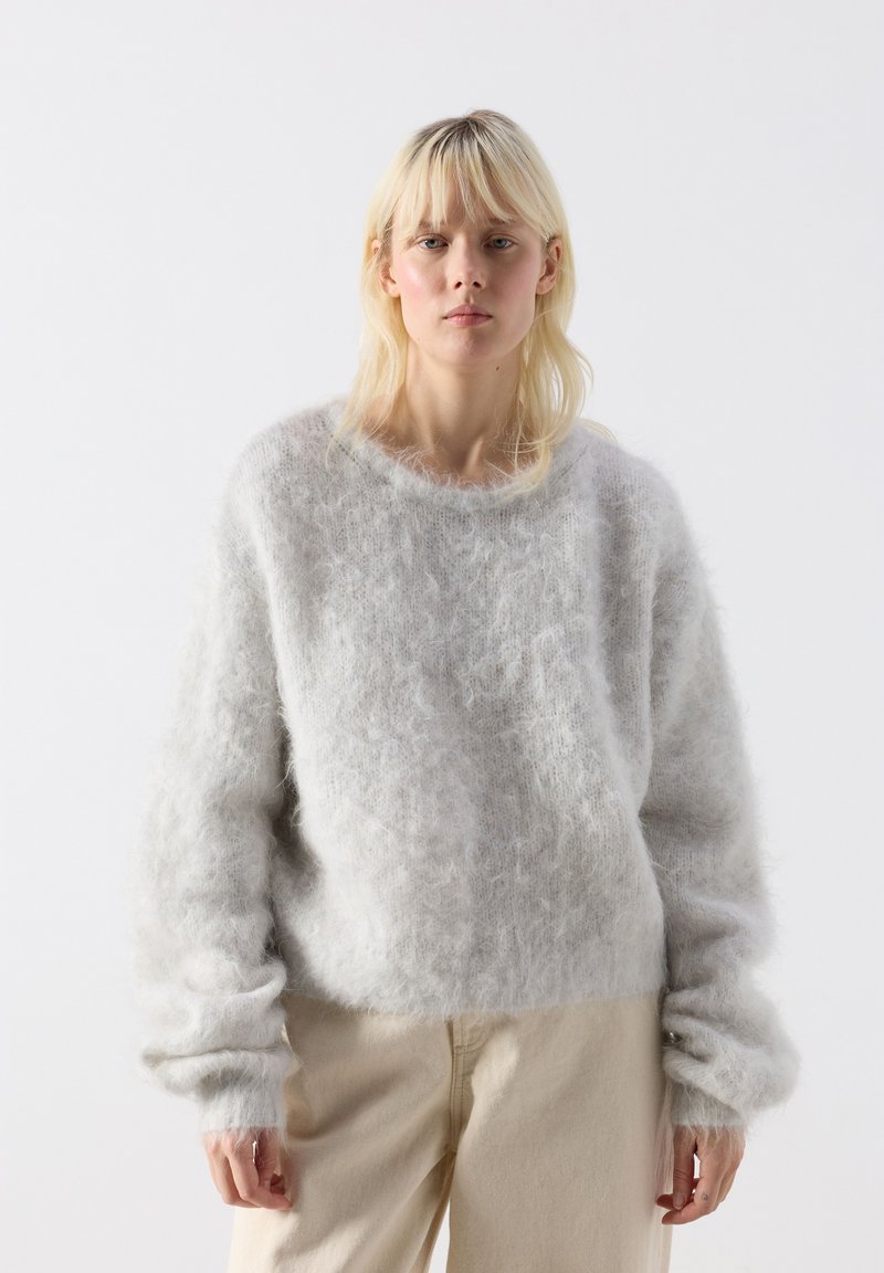 Blonde Frau mit glattem Haar, die einen hellgrauen, flauschigen Pullover und beige Hose trägt, vor einem schlichten weißen Hintergrund stehend.