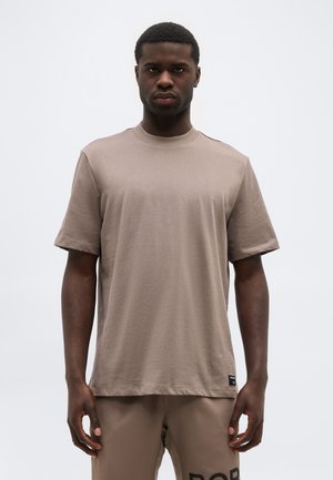 Egy laza, taupe színű pólót és hozzá illő rövidnadrágot viselő férfi áll egy egyszerű, világos háttér előtt.