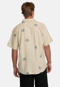 Maglietta a maniche corte in tessuto beige chiaro con un motivo di design circolari verdi e lavanda. La vista posteriore mostra una vestibilità rilassata e un colletto piatto.
