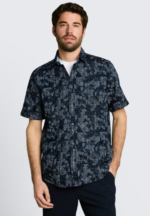 Mężczyzna w granatowej koszuli na krótkim rękawie z abstrakcyjnym jasnoniebieskim wzorem kwiatowym, jedną ręką w kieszeni, stoi na gładkim tle.