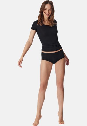 Smilende kvinde stående barfodet iført en tætsiddende sort top med korte ærmer og sorte boyshorts mod en ensartet baggrund.