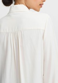 Blouse en crème clair avec un pli au dos, un col à boutons et un tissu doux et lisse. Un éclat subtil améliore la texture et le design.