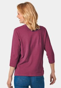 Bordeauxfarbenes T-Shirt mit Dreiviertelärmeln, aus weichem Stoff, mit rundem Ausschnitt und lockerer Passform. Getragen mit blauen Jeans.