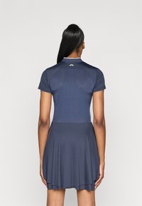 Kobieta z długimi czarnymi włosami w granatowej sukience z krótkim rękawem, z perforowanym tyłem i małym białym logo przy kołnierzyku, odwrócona plecami.
