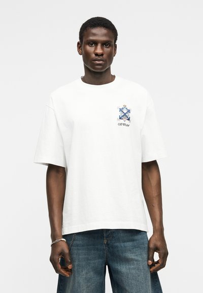 Uomo che indossa una maglietta bianca oversize con emblema blu e dorato e testo "Off-White", abbinata a jeans blu larghi, in piedi davanti a uno sfondo semplice.