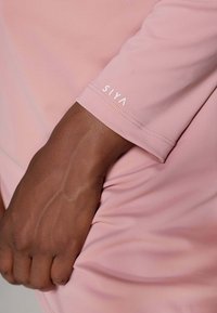 Lichtroze langemouwen shirt met een gladde textuur. Bevat een logo "SIYA" in het wit gedrukt op de manchette. Close-up van de arm en de stof.