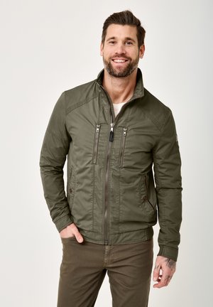Man smiling, wearing zipped green casual jacket and brown pants, standing with one hand in pocket against plain background.