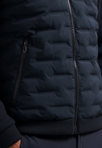 Marineblauwe pufferjas met een gestikte textuur, voorzien van een ritsluiting en ribgebreide boorden. Glad materiaal en slank ontwerp.