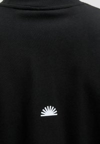 Schwarzes Sweatshirt-Material mit einem weißen Sonnenstrahlen-Logo auf der Rückseite. Der Halsausschnitt ist gerippt und die Textur ist glatt und weich.