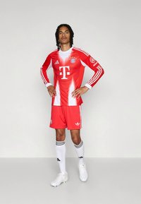 Rotes FC Bayern München Fußballtrikot mit weißen Akzenten, das ein Verlaufsdesign, ein Logo und drei Streifen an Ärmeln und Shorts aufweist.