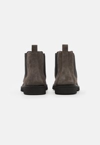 Brązowe zamszowe botki za kostkę z elastycznymi panelami bocznymi, okrągłym noskiem i pętlą do chwytania. Czarna teksturowana gumowa podeszwa zapewnia stabilny chwyt.
