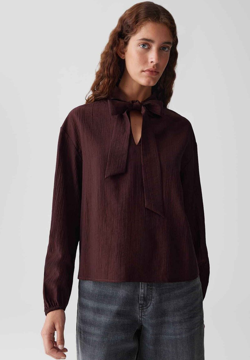 Bordeaux lange mouwen blouse van textuurstof, met een strikaccent bij de hals en een v-vormige opening. Gecombineerd met grijze denim.