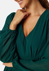Grünes, fließendes Kleid aus leichtem Stoff mit tiefem V-Ausschnitt und voluminösen Ärmeln, die an den Handgelenken für Detail und Dimension gerafft sind.