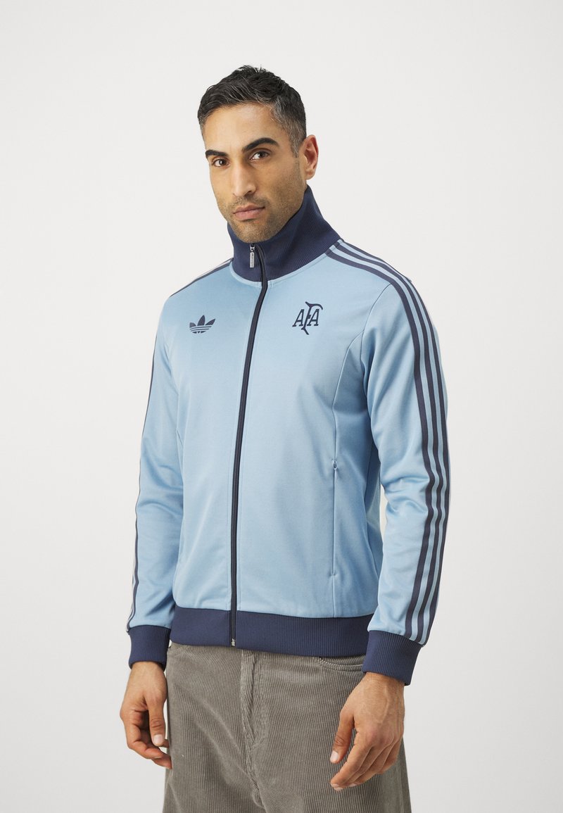 Svijetloplava atletska jakna s visokim ovratnikom, mornarskim akcentima i detaljem s tri pruge. Ima Adidas logotip na prsima i džepove s patentnim zatvaračem.