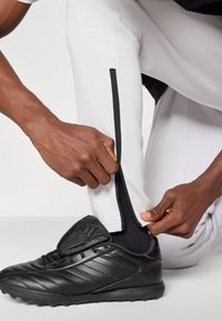 Λευκό αθλητικό παντελόνι με μαύρα φερμουάρ στο πλάι, σε συνδυασμό με μαύρα ποδοσφαιρικά παπούτσια. Το ύφασμα φαίνεται λείο, με εφαρμοστό σχέδιο στους αστραγάλους.