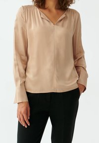 Blusa in satin rosa con scollatura a V, dettaglio arricciato sulle spalle, maniche lunghe e vestibilità comoda, abbinata a pantaloni neri.