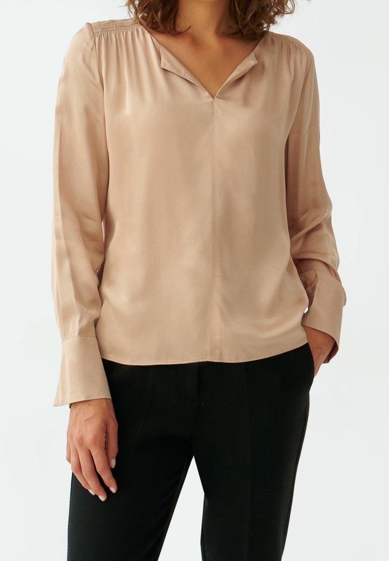 Blusa in satin rosa con scollatura a V, dettaglio arricciato sulle spalle, maniche lunghe e vestibilità comoda, abbinata a pantaloni neri.