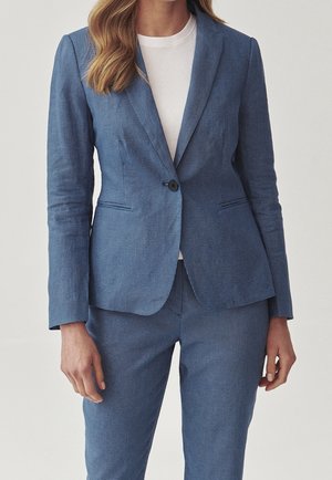 Kvinde iført en tætsiddende blå tekstureret habit med en enkelt-knap blazer over en helt hvid top mod en ensfarvet baggrund.