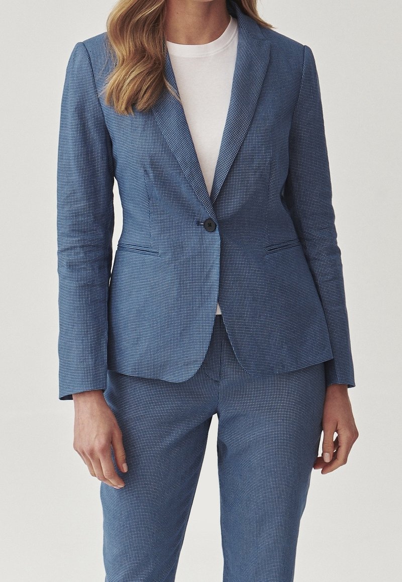Femme portant un tailleur-pantalon bleu ajusté texturé avec un blazer à un seul bouton par-dessus un haut blanc uni sur un fond uni.