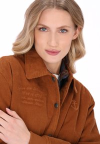 Blonde vrouw met blauwe ogen, gekleed in een bruine ribfluwelen jas, met de armen gekruist, kijkt zelfverzekerd naar de camera tegen een witte achtergrond.