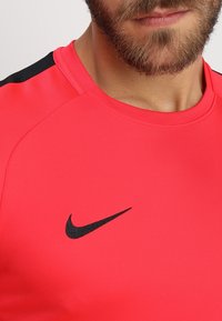 Punainen urheilupaita, jossa on synteettinen materiaali, musta Nike-tunnus ja mustat yksityiskohdat hartioilla.