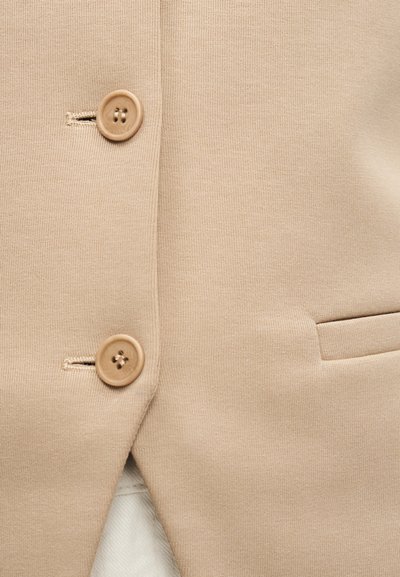 Μπεζ υφασμάτινο σακάκι, μπροστινή όψη με δύο μπεζ κουμπιά και μια οριζόντια τσέπη με άνοιγμα στη δεξιά πλευρά.