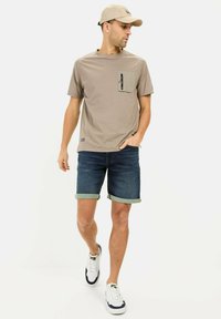 Chemise à manches courtes beige clair avec une poche poitrine et une fermeture éclair noire, associée à un short en denim bleu et des baskets blanches avec des accents marine.