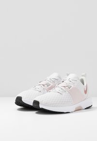 Scarpe sportive bianche con accenti rosa, tomaia texture e suola in gomma nera. Presentano un design con lacci e un tirante sul tallone.