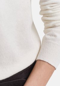 Close-up van een persoon die een zacht wit vest draagt met geribbelde manchetten en een donkerbruin kledingstuk om de taille.