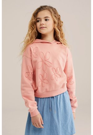 Djevojka s dugom kovrčavom kosom koja nosi ružičastu hoodicu s cvjetnim uzorcima i svijetloplavu suknju, gledajući blago u stranu.