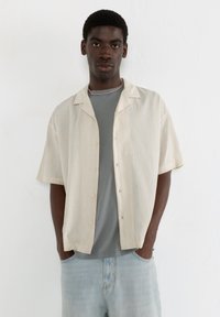 Helle beige kurzärmlige Knopfhemd mit einem strukturierten Muster, getragen über einem grauen T-Shirt, kombiniert mit hellblauen Jeans.