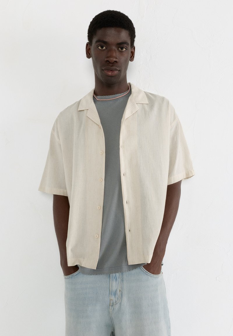 Helle beige kurzärmlige Knopfhemd mit einem strukturierten Muster, getragen über einem grauen T-Shirt, kombiniert mit hellblauen Jeans.