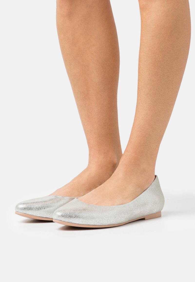Chaussures de ballet plates en argent texturé portées pieds nus, vues de côté sur un fond clair uni.