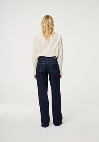 Lichte, gestructureerde beige blouse met lange mouwen, gecombineerd met donkere denim wijde jeans. De jeans hebben twee achterzakken en een label bij de taille.