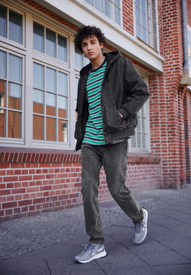 Donkergrijze hoodie over een groen-blauw gestreepte t-shirt, gecombineerd met grijze jeans en lichtgrijze sportschoenen. Stedelijke omgeving.