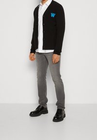 Man som bär en svart knappad kofta med blå "W," vit skjorta, grå jeans och svarta läder snörskor står framför en vit bakgrund.