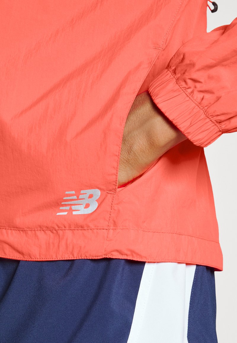 Veste coupe-vent corail avec une finition légère et texturée et des poignets élastiques, ornée d'un logo en métal argenté réfléchissant et d'une ouverture de poche sur le côté.