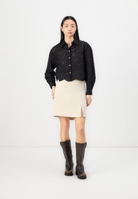Black embroidered button-up shirt with cut-out patterns, paired with a beige ribbed skirt featuring a side slit and brown knee-high boots.