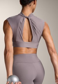 Top corto grigio sportivo con design a nodo sul retro. Tessuto liscio e elastico che offre una silhouette aderente. Il modello tiene in mano una piccola palla di peso grigia.