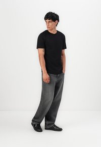 Man standing against white wall wearing black t-shirt, loose gray jeans, and black shoes, looking to his left.