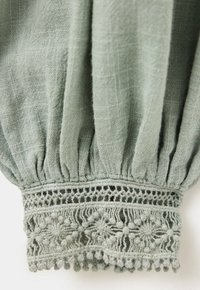 Gros plan d'une manche en tissu vert clair avec un élastique froncé et une bordure en dentelle florale détaillée au poignet.