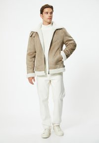 Chaquetón de ante marrón con forro y puños de piel de oveja blanca, que cuenta con una cremallera frontal y dos bolsillos laterales, llevado sobre un suéter blanco y pantalones.