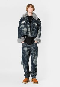 Chaqueta de denim recortada con cuello y puños de piel sintética gris, combinada con pantalones de denim oscuro a juego, ambos con patrones abstractos.