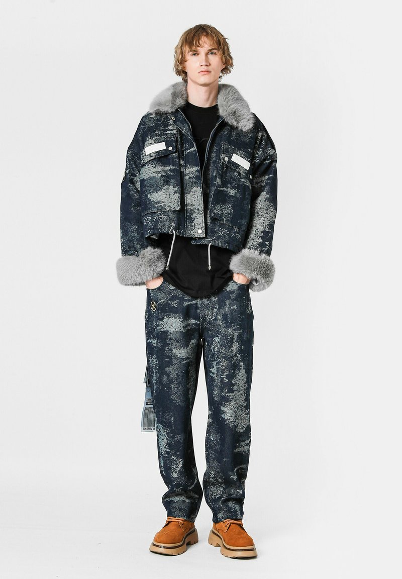 Chaqueta de denim recortada con cuello y puños de piel sintética gris, combinada con pantalones de denim oscuro a juego, ambos con patrones abstractos.