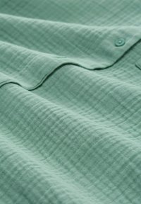 Texturovaná látka v světle zelené barvě, se štepovaným vzorem a detailním knoflíkem. Měkká a hladká textura s jemnými vlnkami.