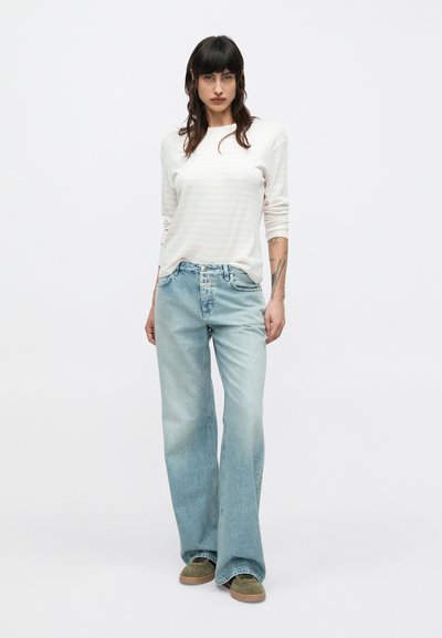 Chemise beige clair à manches longues avec rayures subtiles, associée à un jean évasé bleu clair et des chaussures vert-brun, posant devant un fond neutre.