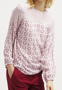 Blusa rosa chiaro a maniche lunghe con un motivo geometrico e dettagli in pizzo sul petto; vestibilità morbida, tessuto morbido e scollatura rotonda.