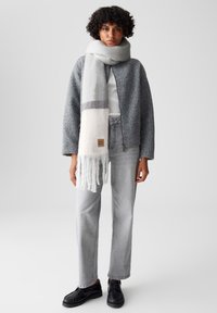 Grijze gebreide jas, witte sjaal met grijze streep, grijze high-waisted jeans en zwarte schoenen. De sjaal heeft franjes aan de uiteinden en een label.